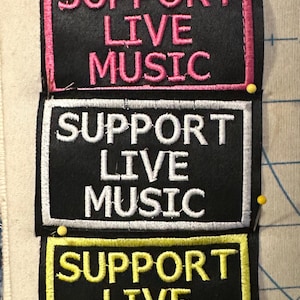 Può includere: Una pila di cinque toppe di tessuto nero con le parole "SUPPORT LIVE MUSIC" ricamate in bianco, rosa e giallo. Ogni toppa ha un bordo colorato ed è fissata con spilli a un tessuto chiaro. Misure non applicabili.