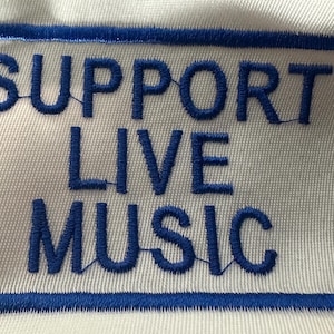 May include: Embroidered patch with the words "SUPPORT LIVE MUSIC" in blue on a white background. The text is enclosed within a blue rectangular border. The patch is likely designed to be sewn onto clothing or accessories.