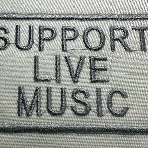 Könnte beinhalten: Ein rechteckiger Aufnäher mit dem gestickten Text "SUPPORT LIVE MUSIC" in dunkelgrauem Faden. Der Text ist innerhalb eines dunkelgrauen Rahmens auf einem hellgrauen Stoffhintergrund zentriert. Der Aufnäher ist wahrscheinlich für Kleidung oder Accessoires.