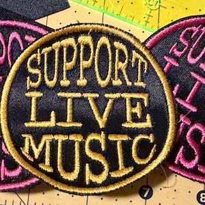 May include: Three circular embroidered patches with black fabric backgrounds. The centre patch features gold thread text reading SUPPORT LIVE MUSIC, while the two flanking patches feature pink thread text reading the same phrase in a circular layout.