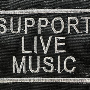 Puede incluir: Parche rectangular negro con las palabras "SUPPORT LIVE MUSIC" bordadas en hilo plateado. El texto está enmarcado por un borde plateado, creando un contraste llamativo sobre un fondo oscuro.