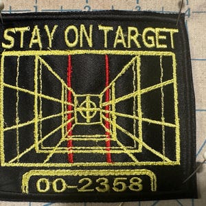 Può includere: Toppa ricamata nera con le parole "STAY ON TARGET" in giallo. La toppa presenta un disegno a bersaglio giallo e rosso con i numeri "00-2358" in basso. La toppa è quadrata e ha un bordo nero.