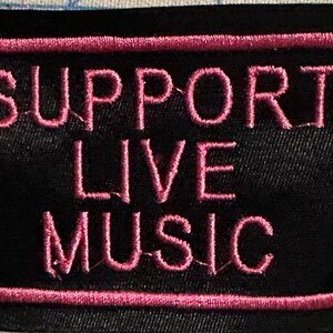 Może przedstawiać: Czarna, haftowana naszywka z różowym napisem "SUPPORT LIVE MUSIC". Tekst jest obramowany różową obwódką. Idealna do odzieży lub akcesoriów.