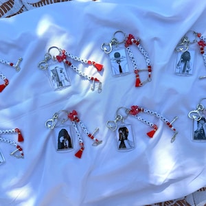 May include: Eight rectangular acrylic keychains featuring black and white portraits of individuals. Each keychain includes a silver metal ring, red and white letter beads spelling ARIRANG and names like NAMJOON, red tassels, and small silver charms.