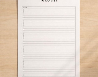 Puede incluir: Un bloc de notas blanco con las palabras "TO DO LIST" impresas en la parte superior. El bloc de notas tiene una cuadrícula de casillas de verificación y espacios con líneas para tareas. El bloc de notas está sobre una superficie de madera marrón claro.