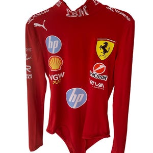Puede incluir: Body rojo de manga larga con múltiples logotipos de patrocinadores, incluyendo HP, Shell y UniCredit. El logotipo de Ferrari está en el pecho. El body tiene un cuello alto y el texto "SKC TECHNOLOGY" está impreso en los hombros.