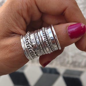 May include: A collection of silver rings with various textures, including braided, etched, and smooth designs. The rings are stacked on a finger, showcasing the different styles. The background is a textured white wall and a black and white geometric floor.