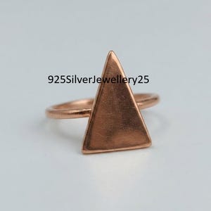 May include: A rose gold-toned ring featuring a prominent, flat, triangular-shaped centerpiece. The metal surface displays fine, visible scratches. The text 925SilverJewellery25 is superimposed over the center of the ring, which has a thin, circular band.