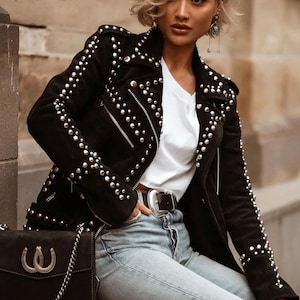 Könnte beinhalten: Schwarze Wildlederjacke mit silbernen Nieten, Reißverschluss und einem weißen T-Shirt darunter. Die Jacke hat einen klassischen Biker-Stil mit Reverskragen und Gürtel. Eine schwarze Handtasche mit silberner Kette ist ebenfalls zu sehen.