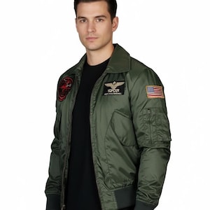 Puede incluir: Chaqueta bomber Top Gun verde oliva con múltiples parches, incluyendo una bandera estadounidense y el texto "TOP GUN". La chaqueta tiene cuello, puños y cintura de canalé, y cierre de cremallera. Tiene dos bolsillos delanteros.