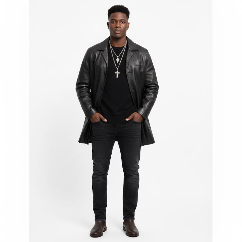 May include: A black leather trench coat with a button-down front, worn over a black t-shirt and dark wash jeans. The outfit is accessorized with a silver cross pendant necklace. The model is wearing brown leather boots.