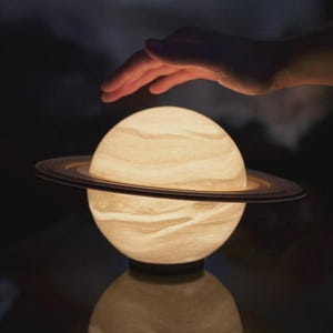 Può includere: Lampada a forma di Saturno, luminosa, con un globo bianco sporco strutturato e un anello marrone scuro. La lampada si riflette su una superficie scura. Una mano fluttua sopra la lampada.