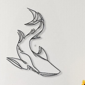May include: Black metal shark wall art. The artwork is a continuous line drawing of a shark, mounted on a white wall. The room also features a wooden table, sunflowers in a gray vase, and black pendant lights.