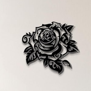 Puede incluir: Una pieza de arte mural de rosa de metal negro. La rosa está intrincadamente cortada con pétalos y hojas detallados. La obra de arte está montada en una pared blanca, agregando un toque de elegancia al espacio. La rosa mide aproximadamente 46 cm de diámetro.