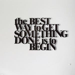 May include: Black metal wall art with the quote "the BEST WAY to GET SOMETHING DONE is to BEGIN" on a white wall. A white staircase and a white vase with branches are also visible.