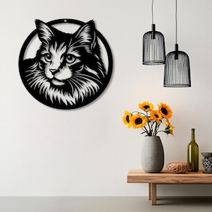 Könnte beinhalten: Schwarze Metall-Wandkunst mit einem Katzenkopf. Das Gesicht der Katze ist mit weißem und schwarzem Fell detailliert, das in einem schwarzen kreisförmigen Rahmen eingefasst ist. Das Kunstwerk ist an einer weißen Wand angebracht, mit Sonnenblumen in einer Vase und hängenden schwarzen Lampen im Hintergrund.