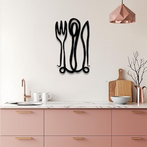 Op de afbeelding: Zwarte metalen wandkunst met een vork, lepel en mes in een doorlopend lijnontwerp. De keukendecoratie is gemonteerd op een witte muur boven een roze aanrecht met gouden handgrepen. Een koperkleurige hanglamp hangt erboven.