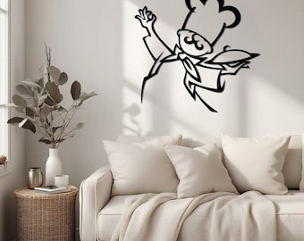 May include: A black metal wall art piece depicting a chef in mid-air, holding a covered dish and making an 'okay' gesture. The chef is wearing a tall hat, a bow tie, and a jacket. The art is mounted on a white wall above a cream-colored sofa.