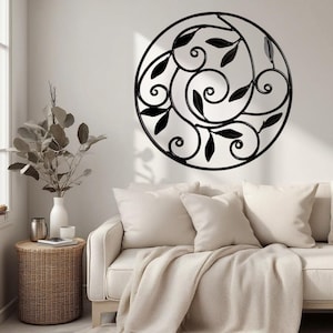 May include: A black metal wall art piece with a circular design. The art features swirling lines and leaf-shaped accents. The wall art is mounted on a white wall above a cream-colored sofa with pillows and a blanket. A vase with decorative branches sits on a side table.