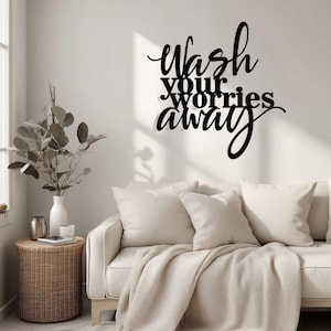 May include: Black metal wall art featuring the words "Wash your worries away" in a cursive script. The artwork is mounted on a white wall above a cream sofa with cushions and a throw. A wicker side table and a vase with foliage are also in view.