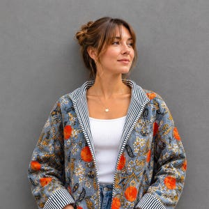 May include: A quilted jacket featuring a blue background with an orange fruit and white floral pattern. The jacket has long sleeves with black and white striped cuffs, a matching striped collar, and two front pockets. It is worn over a white top.