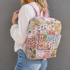 Peut inclure: Un sac à dos matelassé présentant un motif floral patchwork coloré dans des tons de rose, orange, vert et jaune. Le sac comprend une poche avant avec fermeture à rabat, une poignée supérieure et des bretelles réglables avec bordure rayée rose.