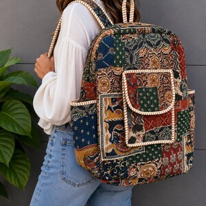 Op de afbeelding: Een gewatteerde rugzak met een veelkleurig patchworkontwerp met ingewikkelde bloemen- en geometrische patronen in tinten dieprood, bosgroen, marineblauw en goud. De tas is voorzien van een voorvak met klep en zijvakken met gestreepte randen.