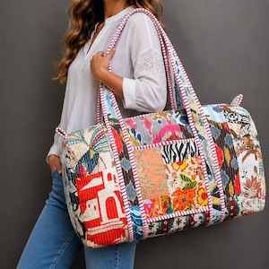 Op de afbeelding: Een grote, rechthoekige reistas van doorgestikte stof met een levendige patchwork van veelkleurige bloemen-, geometrische en dierenprints. De tas heeft twee lange schouderbanden met een rood-witte gestreepte bies.