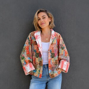 May include: A long-sleeve jacket featuring a multi-colored patchwork pattern with red, orange, and blue tones, worn over a white tank top with light blue denim jeans and brown leather sandals. The jacket has wide cuffs and a white interior lining.