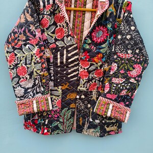 May include: A quilted jacket featuring a dark navy blue base with a vibrant patchwork of floral, botanical, and tiger motifs in shades of pink, red, green, and white. The garment has long sleeves with folded cuffs and a striped trim along the edges.