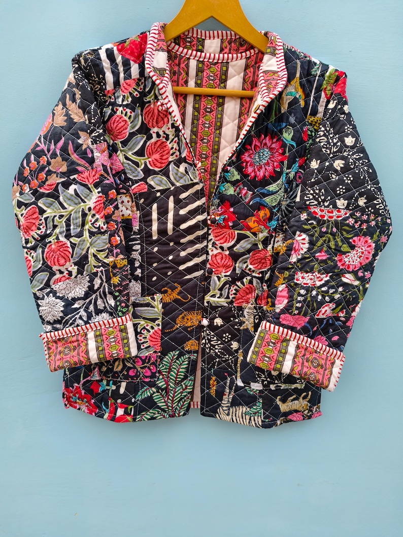 May include: A quilted jacket featuring a dark navy blue base with a vibrant patchwork of floral, botanical, and tiger motifs in shades of pink, red, green, and white. The garment has long sleeves with folded cuffs and a striped trim along the edges.