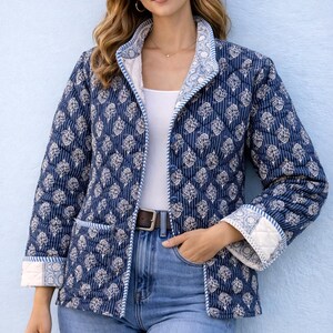 May include: Navy blue quilted jacket with a white floral pattern and vertical stripes. The jacket has a white lining and trim, worn with blue jeans and a brown belt. The jacket is buttoned and has pockets. The jacket's length is approximately 60 cm.