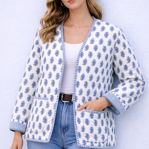 Può includere: Giacca bianca con stampa paisley blu e bordi a righe. La giacca aperta è indossata con un top bianco e jeans blu. È visibile una cintura marrone.