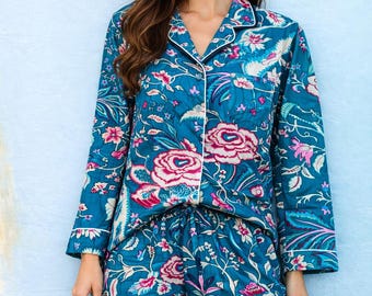 May include: A long-sleeved button-down pajama top and matching pajama pants set in a deep teal blue fabric. The garment features a vibrant floral pattern in shades of pink, white, and light blue, accented with white piping along the collar and cuffs.