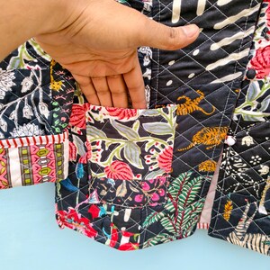 May include: A quilted jacket featuring a patchwork design with black, white, red, and green floral and tiger patterns. The garment includes a front pocket, diamond-shaped stitching, and white buttons. The fabric displays a mix of botanical and animal motifs.