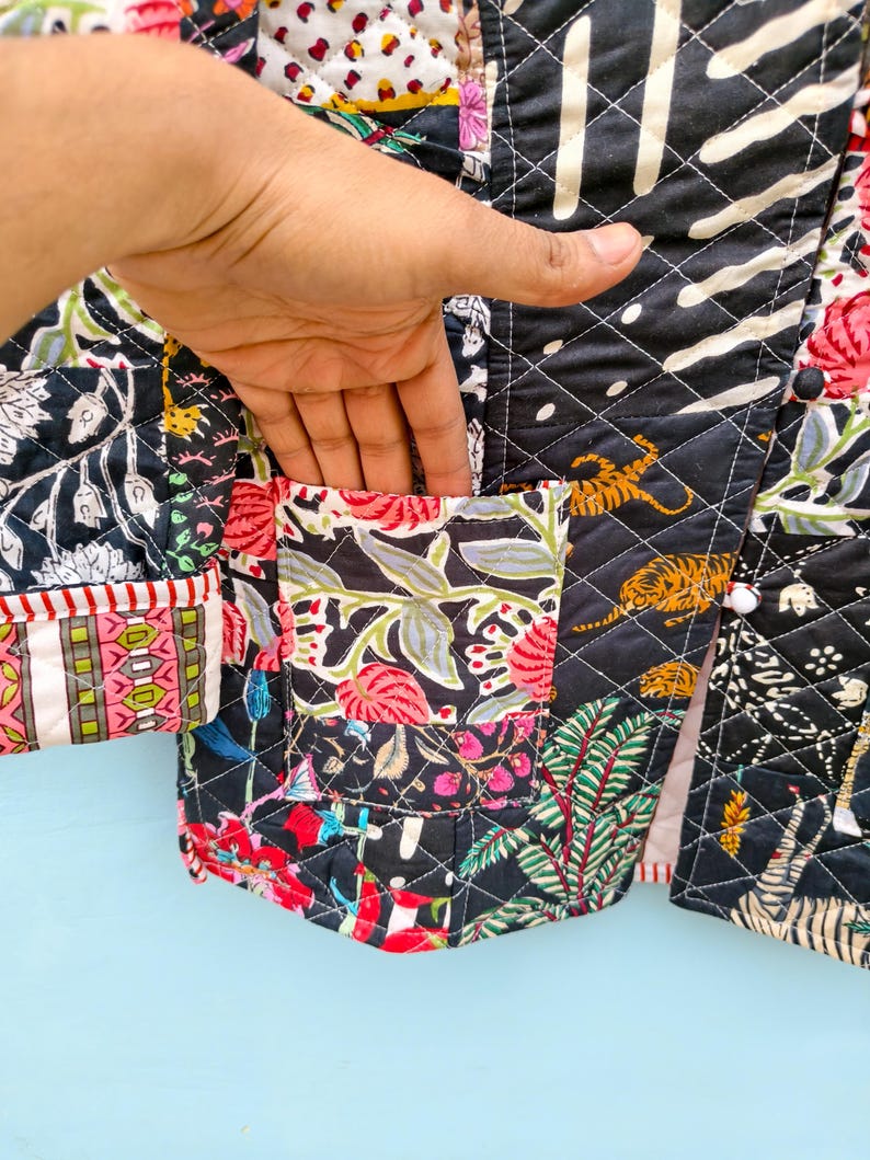 May include: A quilted jacket featuring a patchwork design with black, white, red, and green floral and tiger patterns. The garment includes a front pocket, diamond-shaped stitching, and white buttons. The fabric displays a mix of botanical and animal motifs.