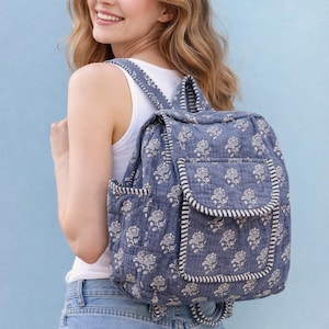 Puede incluir: Una mochila azul con estampado floral, ribete blanco y un bolsillo con solapa. La mochila tiene un diseño acolchado y correas ajustables. La bolsa está hecha de un material suave y ligero, perfecta para llevar libros o artículos personales.