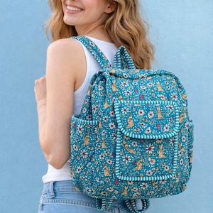 May include: A teal backpack with a floral and rabbit pattern. The backpack has a flap closure, side pockets, and padded straps. The design includes white flowers, red accents, and a striped border. The backpack is made of quilted fabric.