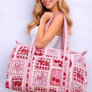 May include: A pink and white quilted duffel bag with a repeating pattern of cherries, bows, and checkered squares. The bag has pink striped handles and trim. The bag is approximately 20 inches long.