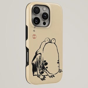 May include: A beige phone case featuring a black and white illustration of a frog. The frog has large eyes and is sitting with its legs spread. Japanese text and a red seal are at the top.