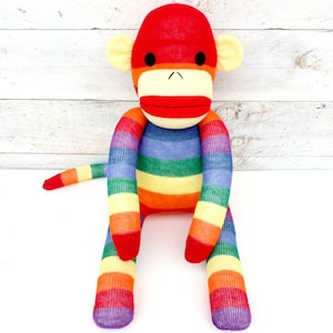 May include: A colourful sock monkey toy with a red head, cream ears, and a rainbow striped body. The monkey has black eyes, a red mouth, and a tail. The doll is made from socks and is a traditional toy.