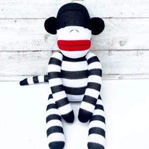 May include: A sock monkey toy with a black and white striped body, black head and ears, and a red mouth. The monkey is sitting upright with its arms and legs extended.