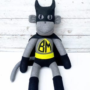 May include: A grey sock monkey dressed as a superhero. The monkey is wearing a black cape, a yellow belt, and a black mask with the letters "BM" on the chest.