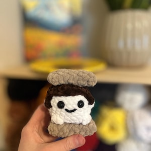 以下が含まれることがあります： 笑顔の顔をした手作りのかぎ針編みのスモアのおもちゃ。スモアは、白いマシュマロ、茶色のチョコレート層、2つのタン色のグラハムクラッカー部分が特徴です。おもちゃは、ぼやけた背景に対して手で持たれています。