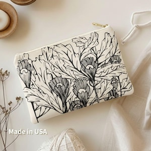 Könnte beinhalten: Cremefarbene Reißverschlusstasche mit schwarzem Blumendesign. Die Tasche hat einen goldfarbenen Reißverschluss und besteht aus einem leinwandartigen Material. Das Blumenmuster umfasst detaillierte Blätter und Blütenknospen. Der Text "Made in USA" ist sichtbar.