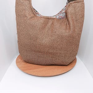 Puede incluir: Un bolso hobo de tela tejida marrón con un asa a cuadros. La bolsa está sobre una bandeja redonda de madera. El forro interior de la bolsa tiene un diseño floral.