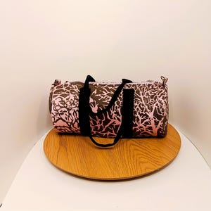 Könnte beinhalten: Rosa-braune Reisetasche mit einem Baumzweigmuster. Die Tasche hat schwarze Griffe und einen schwarzen Riemen. Die Tasche liegt auf einer runden Holzoberfläche.