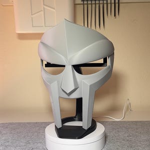 Puede incluir: Una máscara gris angular con un diseño distintivo, montada sobre un soporte negro encima de una base giratoria blanca. La máscara tiene una cara estilizada con rasgos prominentes. El fondo incluye herramientas y una panel perforado.