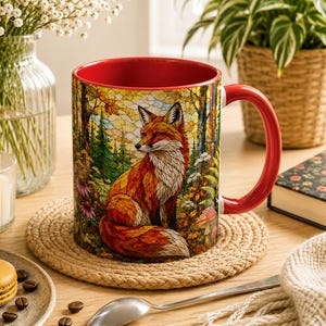 Peut inclure: Un mug en céramique avec un intérieur et une anse rouge vif, orné d'une illustration détaillée et colorée d'un renard assis dans une forêt avec un motif de style vitrail. Le mug repose sur un dessous de verre rond, tissé et beige.