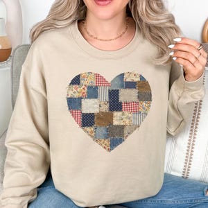 Peut inclure: Un sweat-shirt beige orné d'un cœur en patchwork. Le cœur est composé de divers carrés de tissu bleus, rouges et bruns. Le sweat-shirt a des manches longues et un col rond.